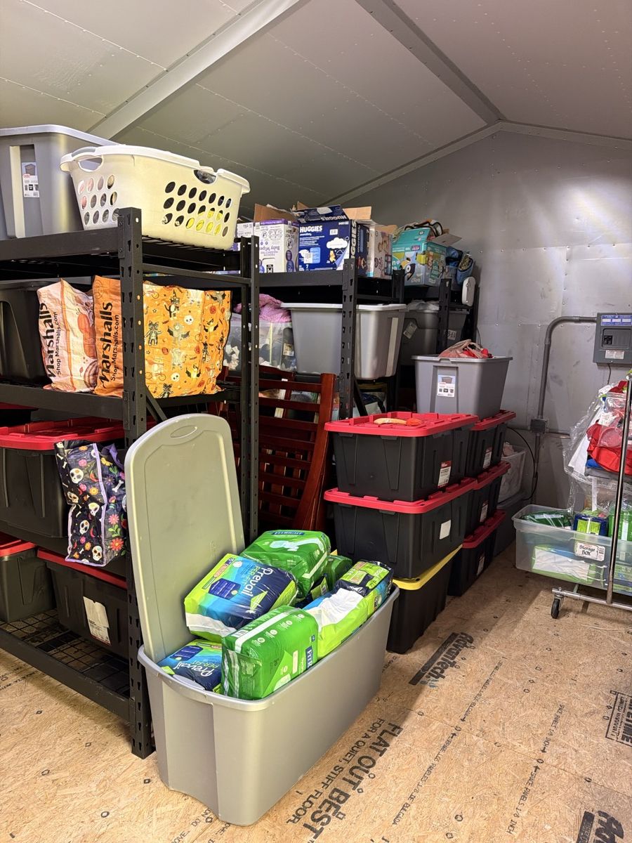 Warehouse shelves with red-lidded storage bins, laundry baskets, packs of adult briefs, and a wooden crib.