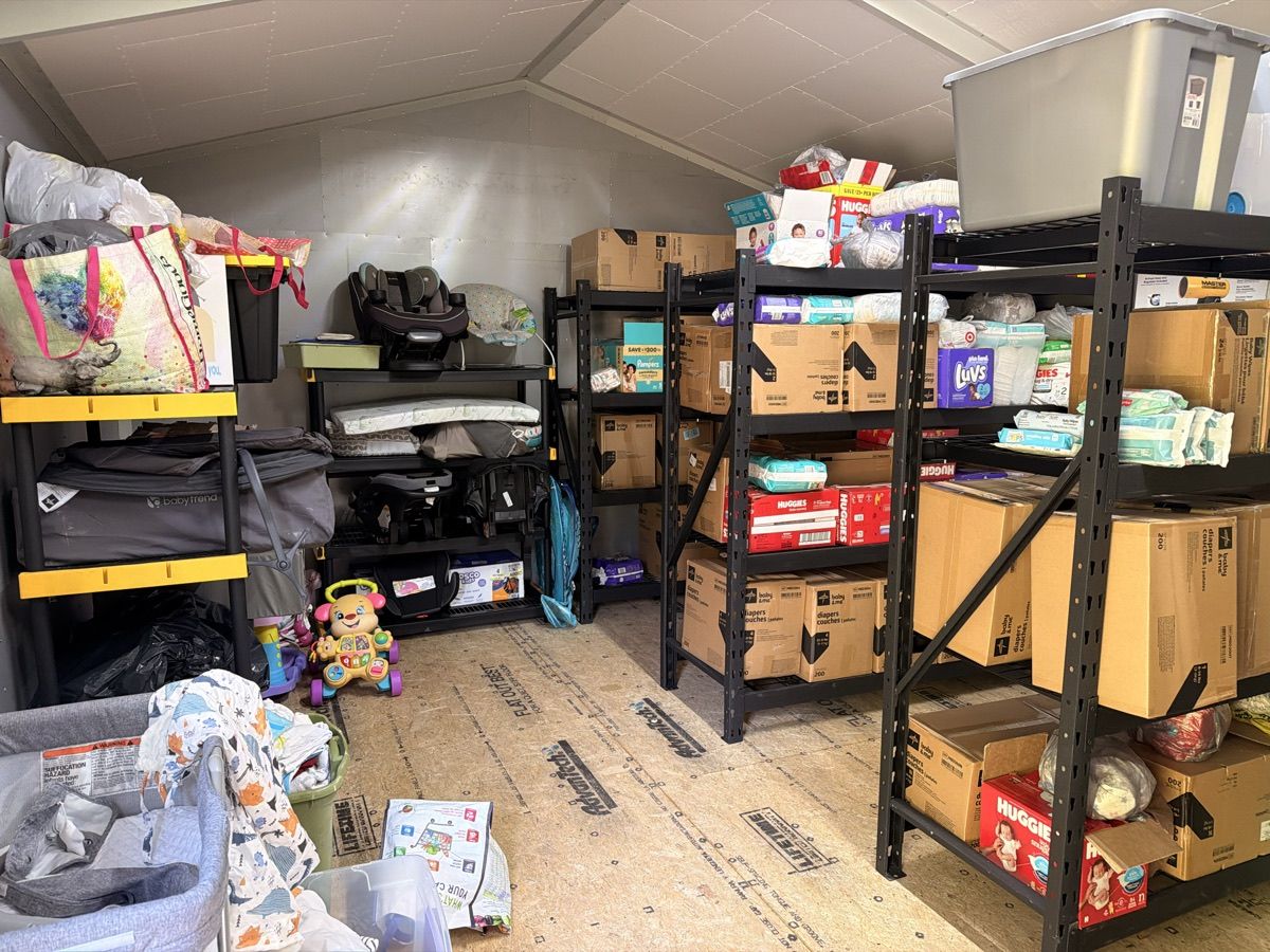 Inside the new warehouse: metal shelves stacked with boxes of Huggies and Luvs diapers and packs of wipes, with car seats, a baby walker, and bags of clothing nearby.