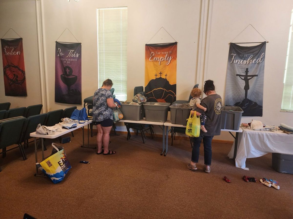 Two moms choosing free clothing and diapers from bins on tables at the Baby Pantry, one holding her toddler on her hip.